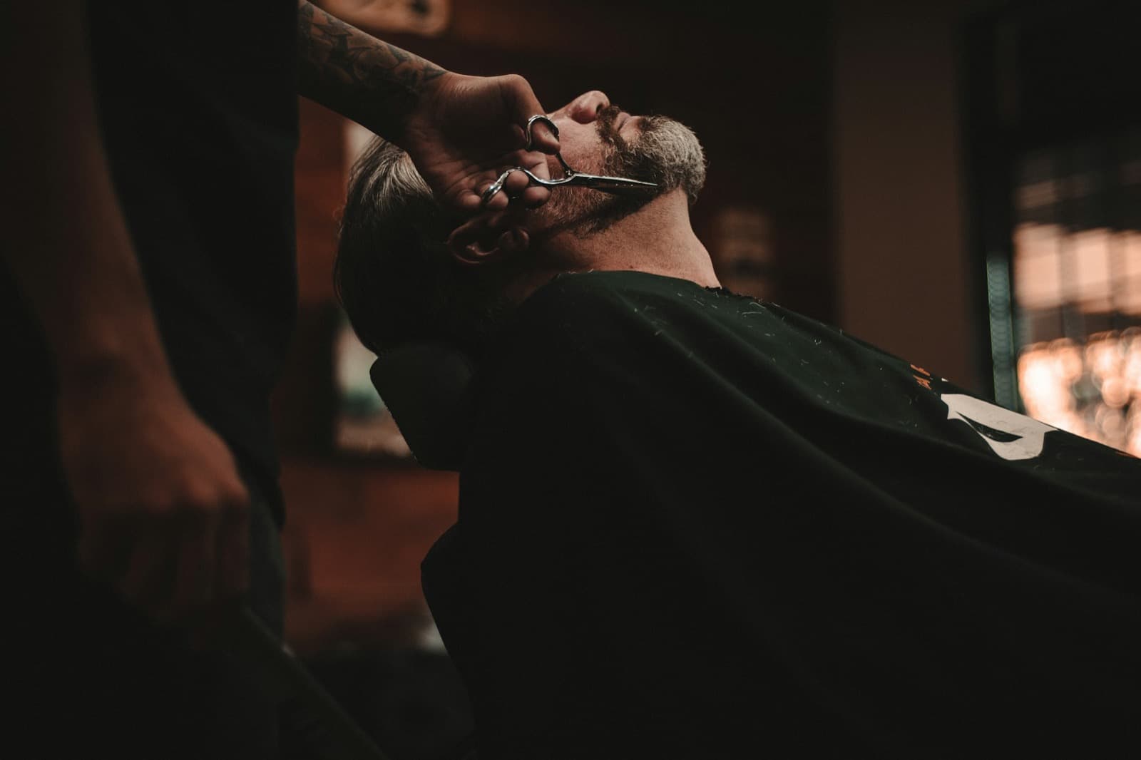 A barber trimming a client's beard with scissors in a warm, dimly lit barbershop